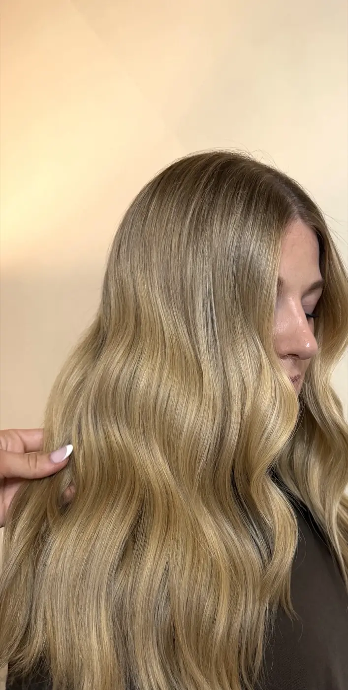 Warm golden blonde waves against a cream backdrop, the client’s face partly turned away.